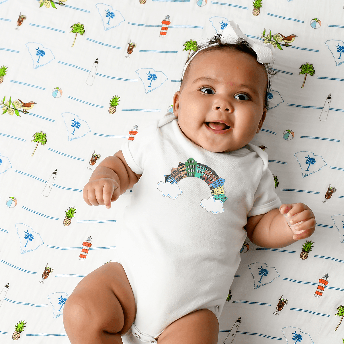 A baby in a white bow lies smiling on a swaddle, wearing the Rainbow Row Charleston baby onesie.