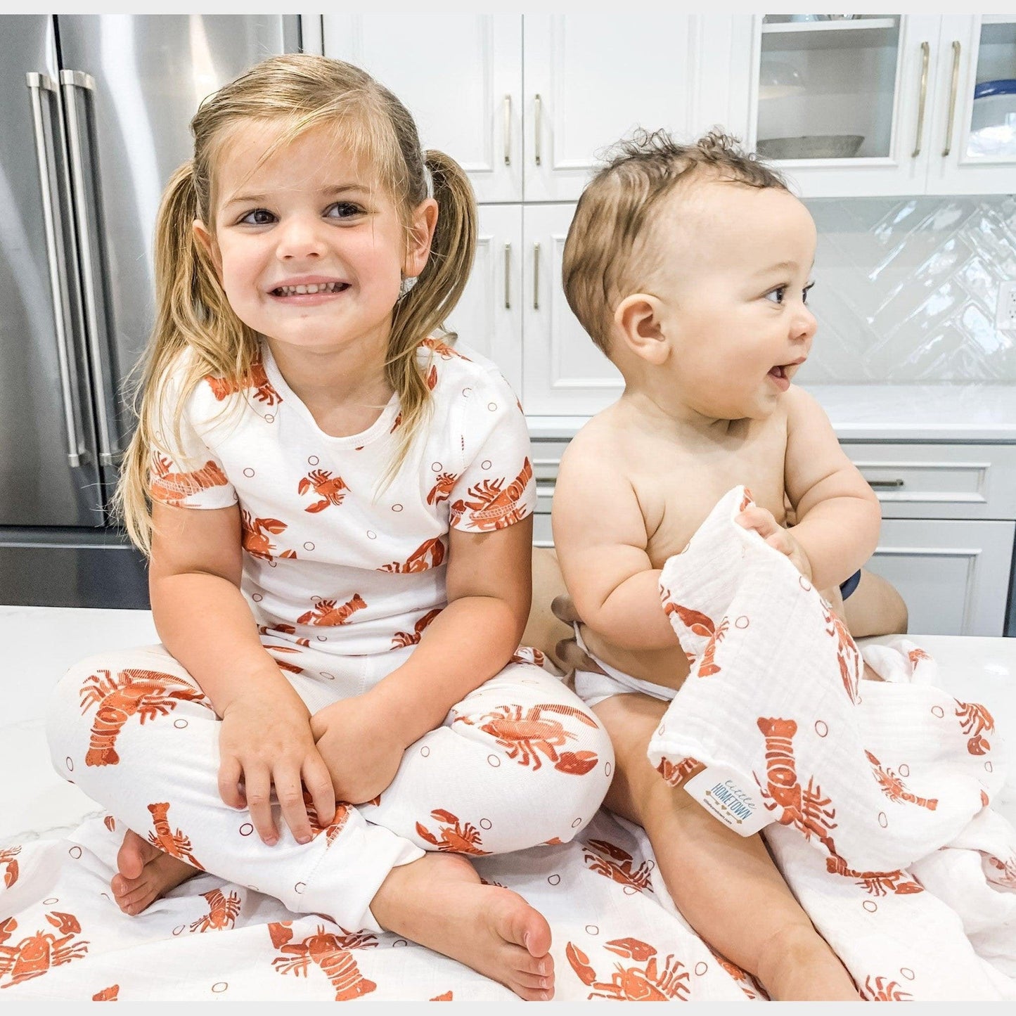 A big sister and her baby brother sit together on the kitchen floor in matching Heads or Tails baby pajamas.