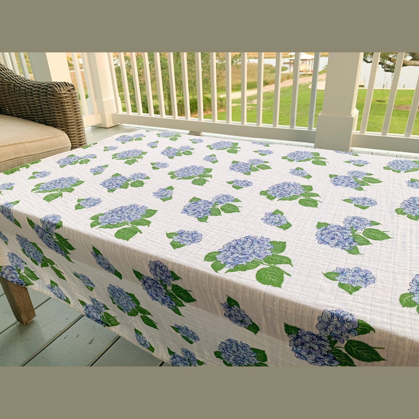 The Hydrangeas muslin swaddle blanket spread over a porch table with the railing and greenery behind it.