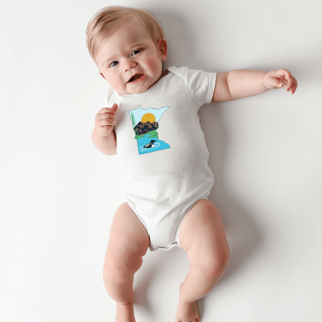 A smiling baby stretches out in the Minnesota Loon baby onesie, resting on a soft neutral blanket.