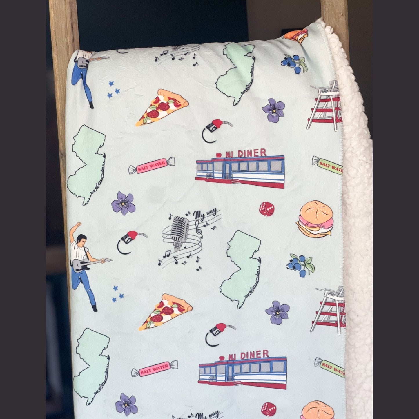 New Jersey-themed plush throw blanket, 60x80 inches, featuring a detailed map with landmarks, a rock star with his guitar, pizza, a diner, a gas pump, musical notes, flowers, and vibrant colors.