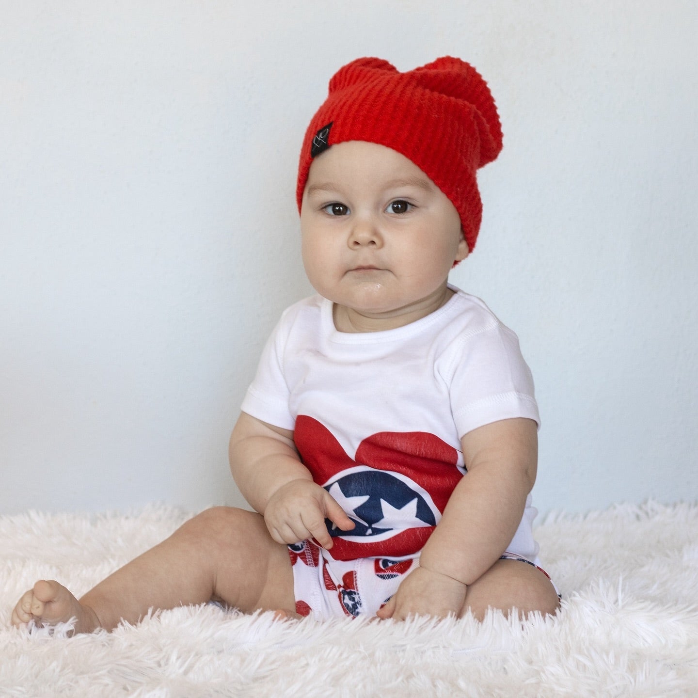 A baby in a red knit hat sits on a fluffy rug wearing the Tennessee Heart baby onesie, all smiles.