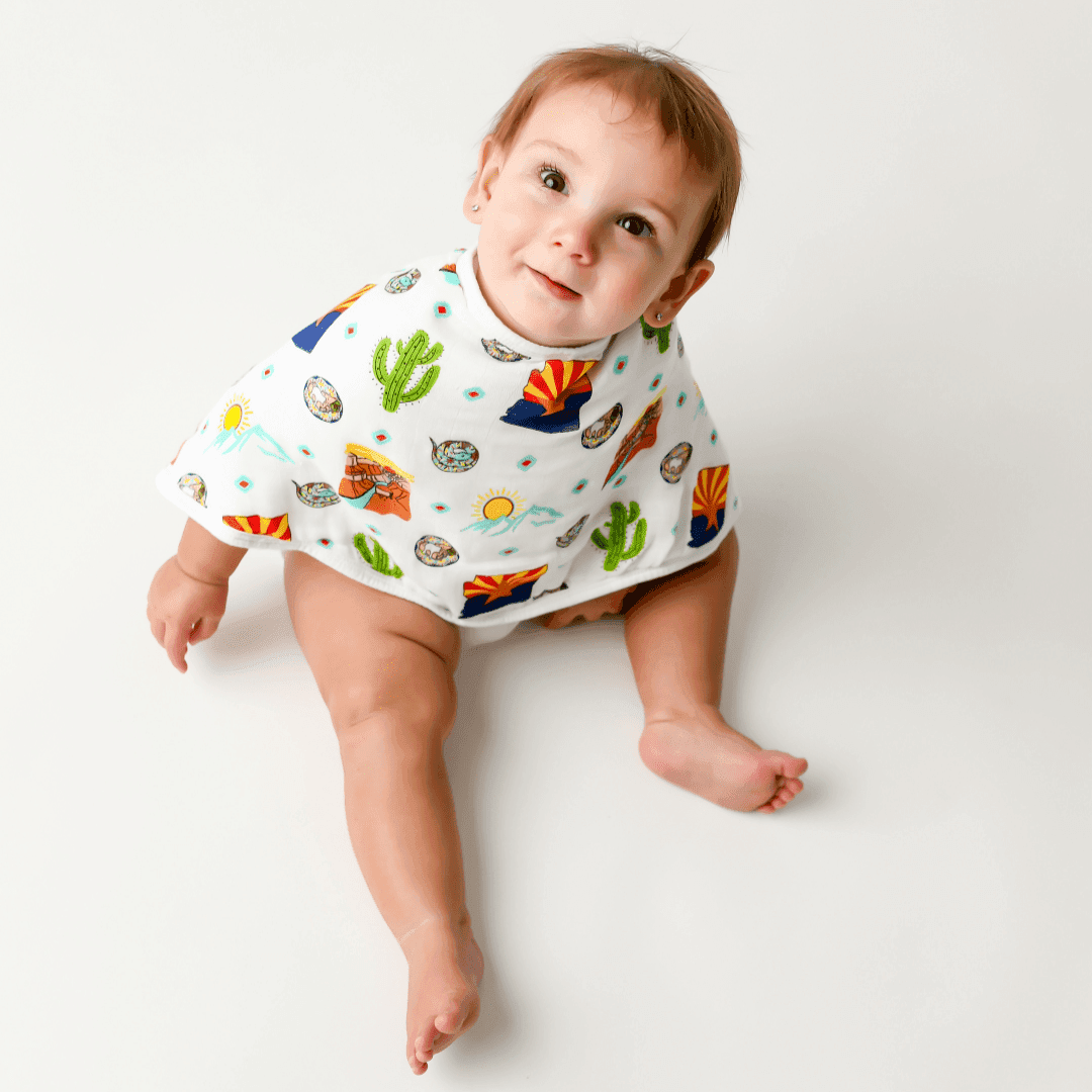 A baby sits up in the Arizona burp cloth and bib combo, looking straight up with round curious eyes.