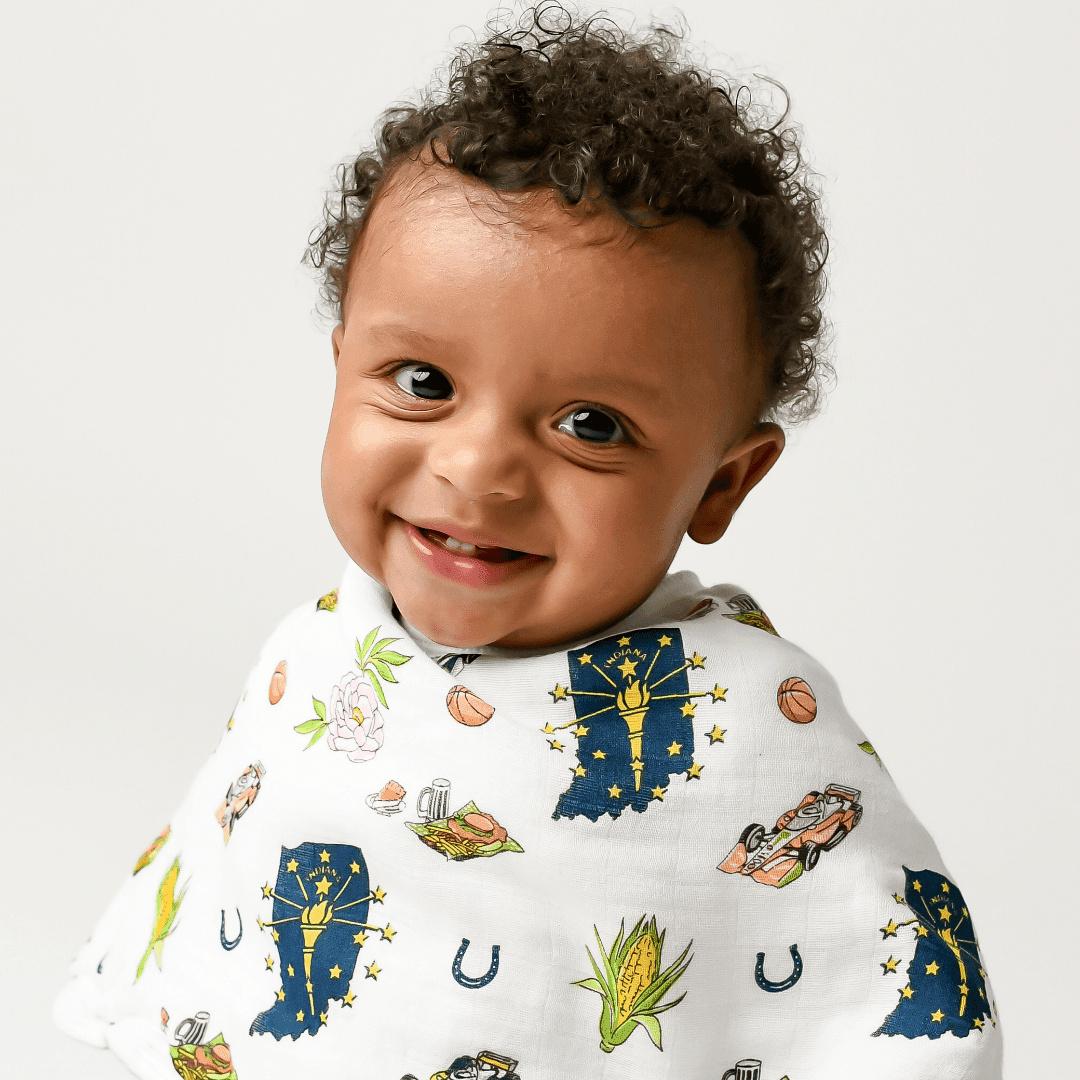 A baby grins from a soft studio backdrop, dressed in the Indiana burp cloth and bib combo.