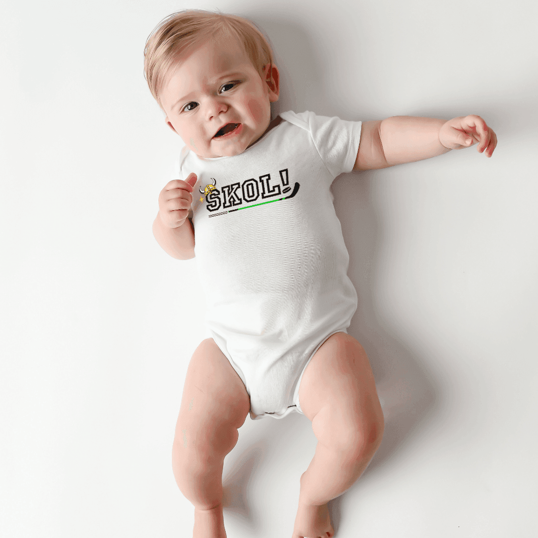 A baby lies smiling in the Minnesota Skol baby onesie, stretched across a soft neutral surface.