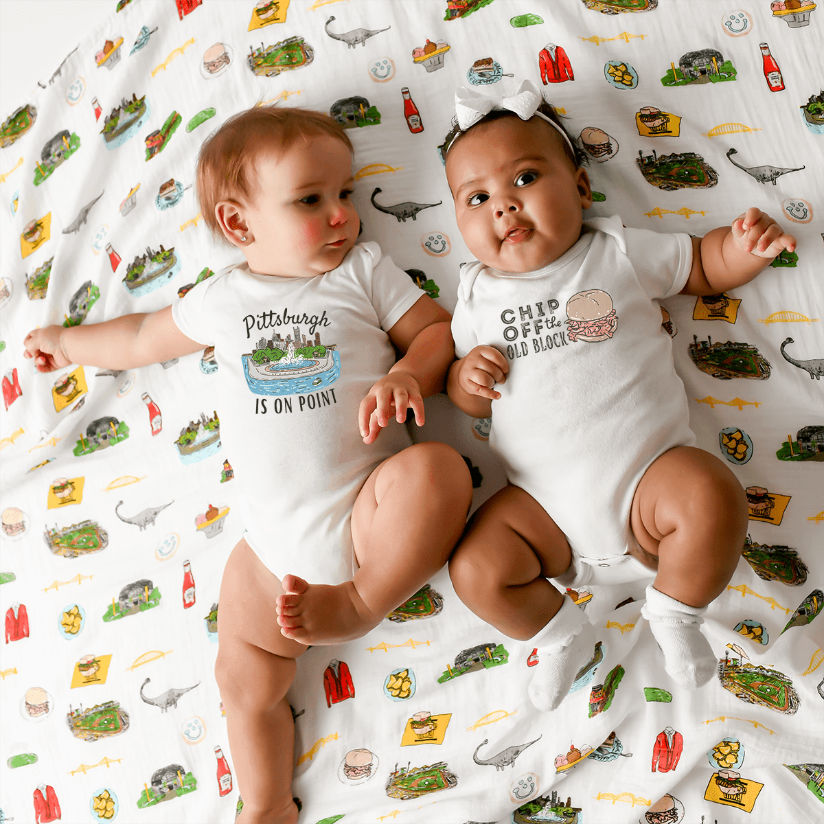 Two babies stretch out together on a Pittsburgh swaddle, matching in the is on Point baby onesie.