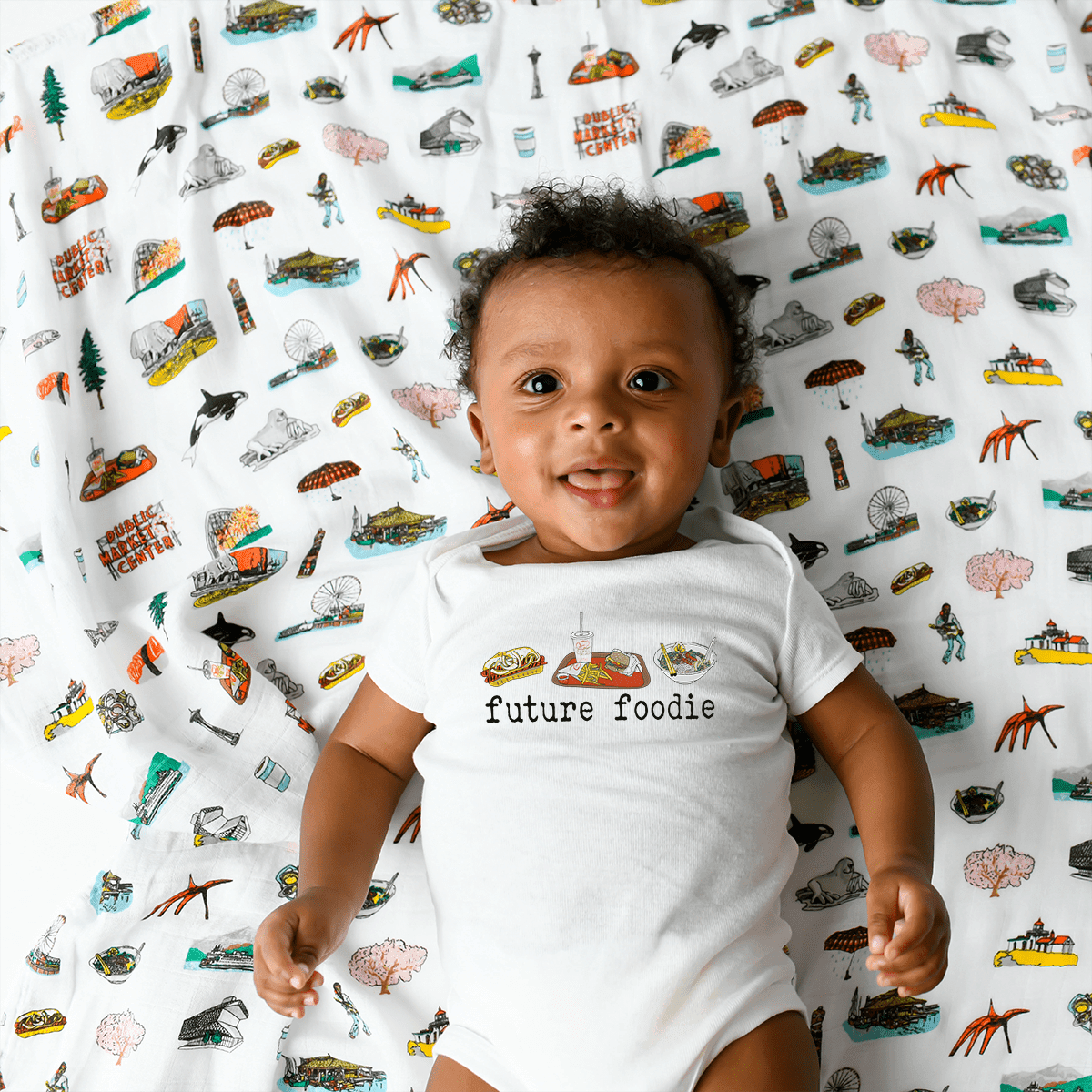 A grinning baby in the Seattle Foodie baby onesie lies back on a city print swaddle in the crib.