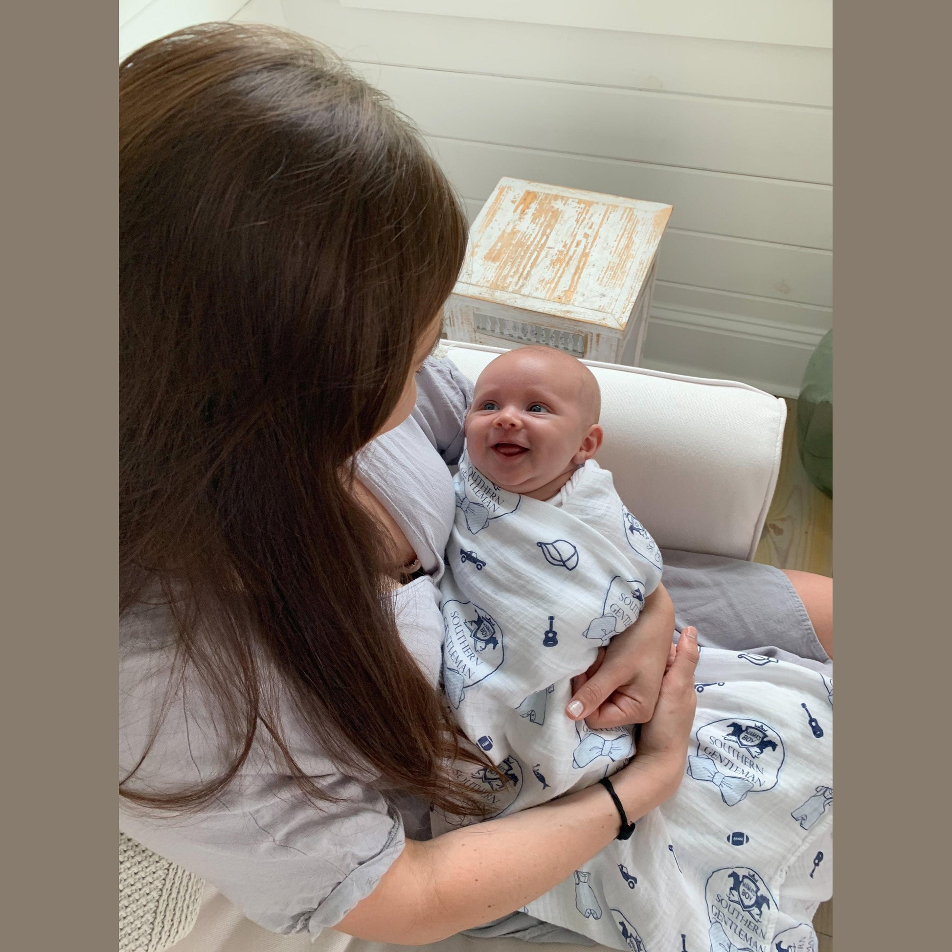 Baby boy swaddled in a white muslin blanket with blue bow ties and mustaches, lying on a soft white surface.
