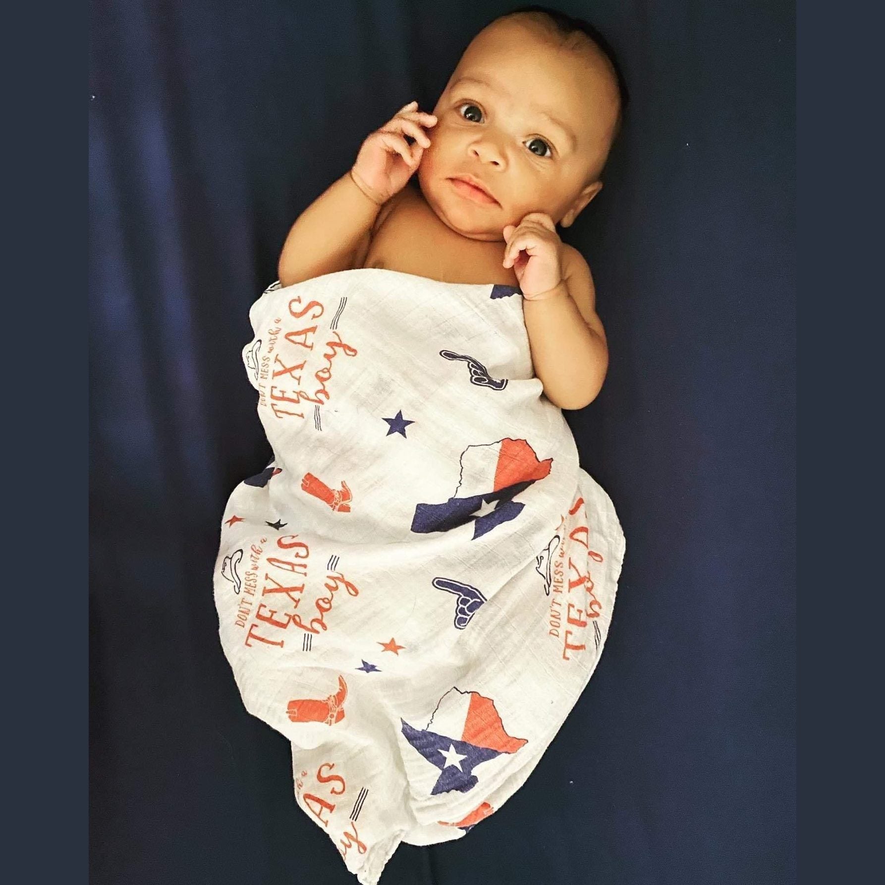 White muslin swaddle blanket with blue Texas-themed illustrations, including the state outline, cowboy boots, and stars.