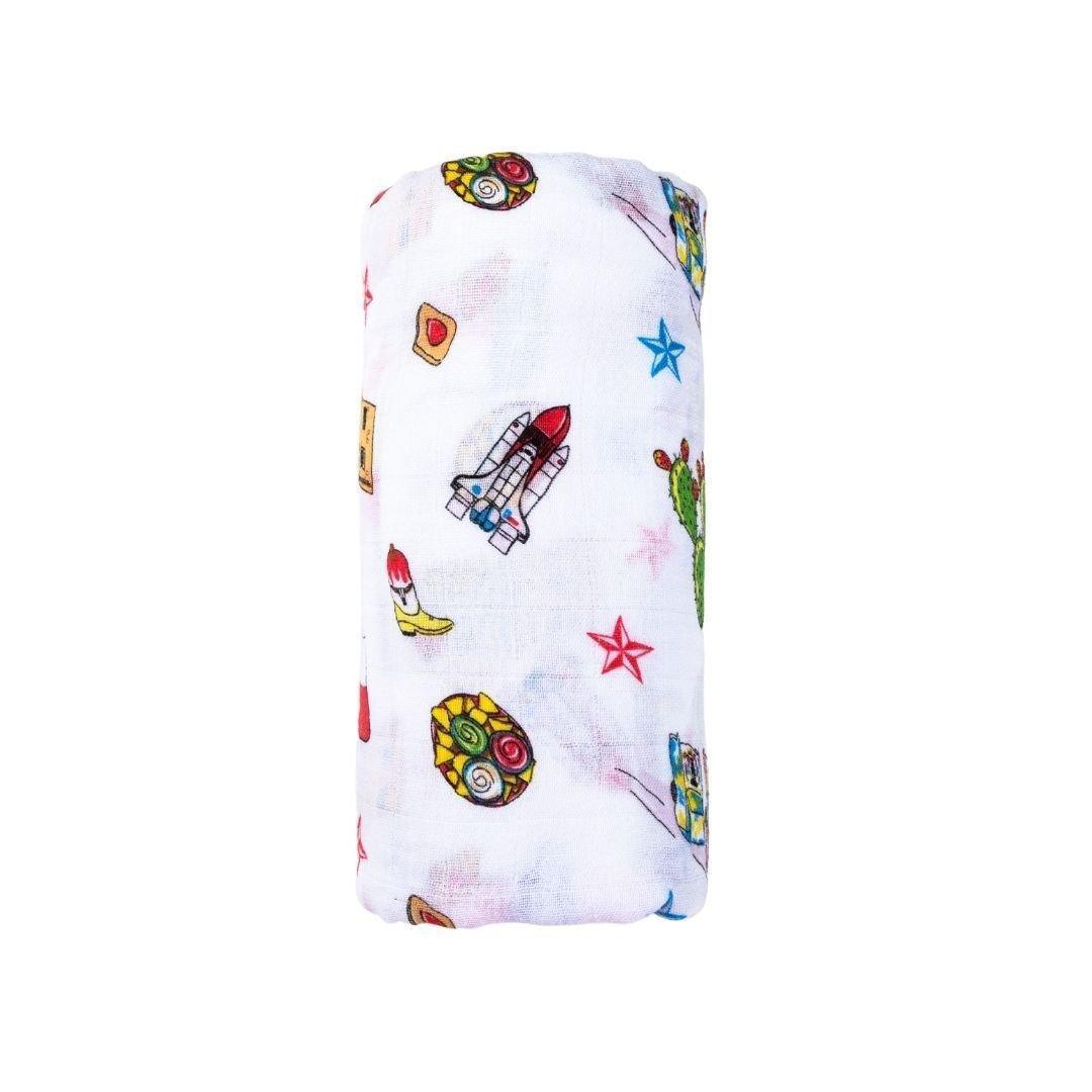 White muslin swaddle blanket with colorful Texas-themed illustrations, including cacti, cowboy boots, and armadillos.