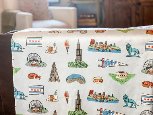 Cozy Chicago-themed plush throw blanket, 60x80 inches, featuring iconic city landmarks like deep dish, a Ferris wheel, lion, bean, a boat on a green river, Windy City flag, and other elements in vibrant colors.