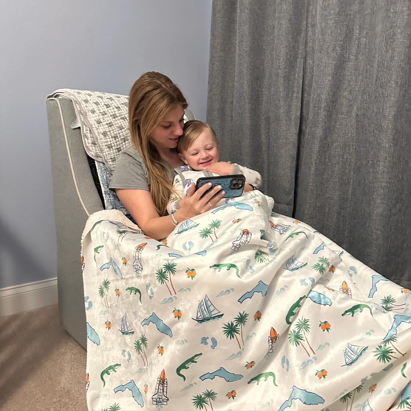 A mom and smiling child cuddling under a Florida-themed plush throw blanket, 60x80 inches, featuring vibrant tropical designs, oranges, alligators, manatees, palm trees, space shuttle, and other Florida elements