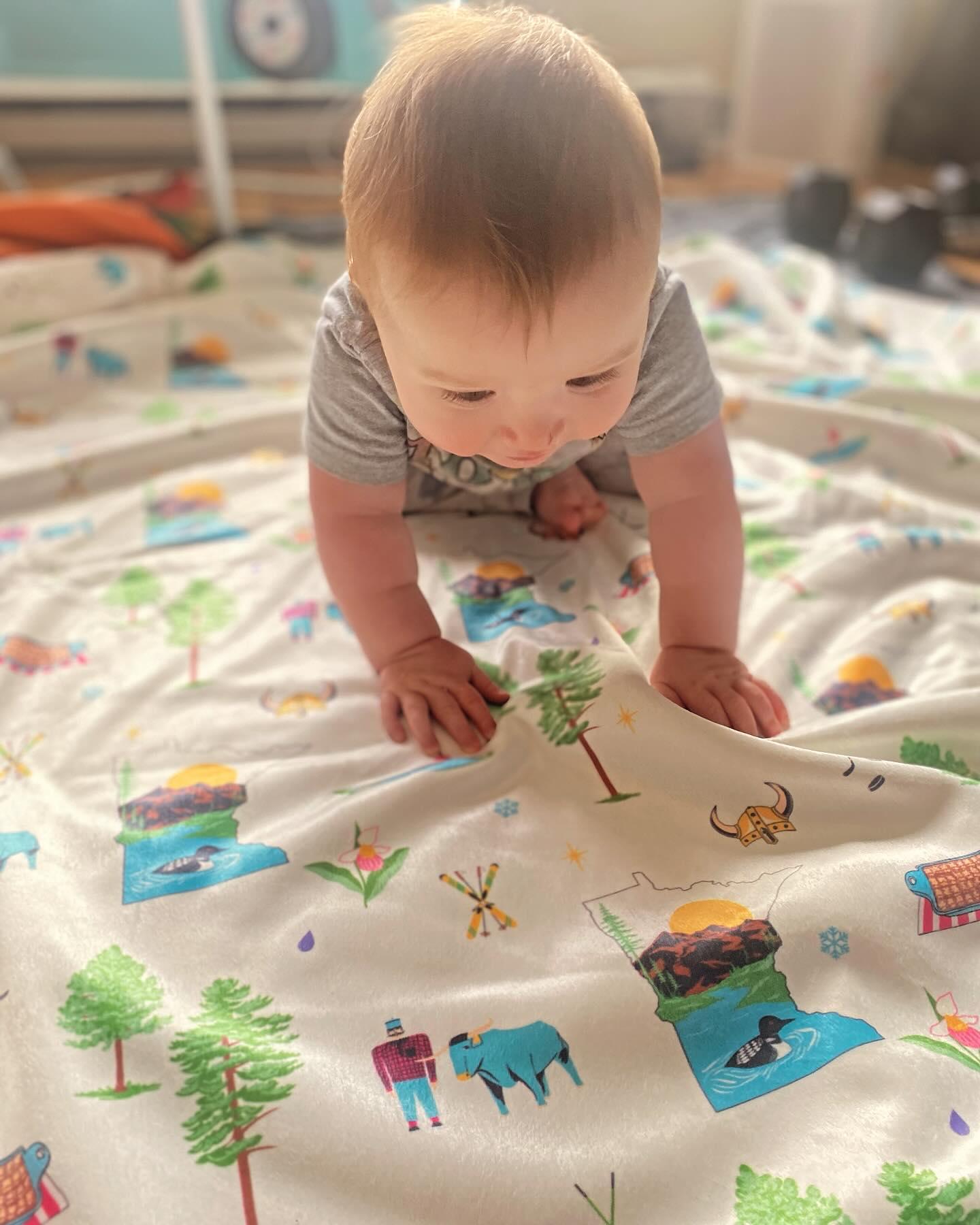 A baby crawling over a Minnesota-themed muslin swaddle blanket, featuring state icons and an outline of the state map