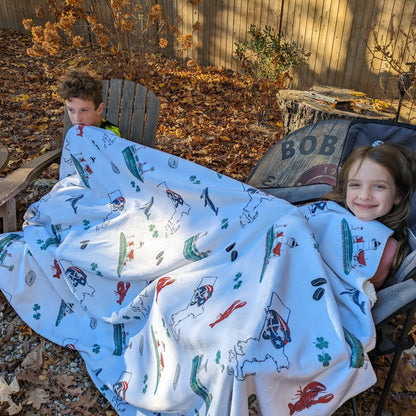 Boy and girl cuddling outside on a cool Fall day both sharing a Massachusetts-themed plush throw blanket, 60x80 inches, featuring a detailed map with landmarks and vibrant colors.