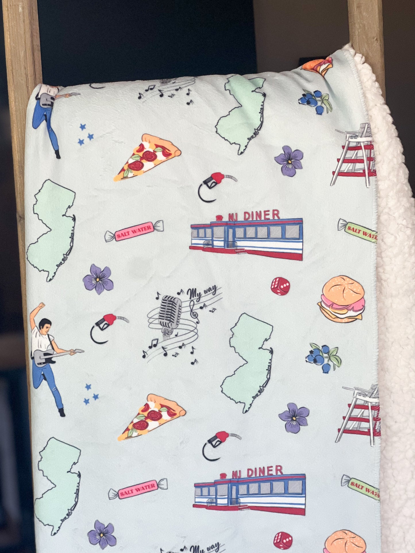 New Jersey-themed plush throw blanket, 60x80 inches, featuring a detailed map with landmarks, a rock star with his guitar, pizza, a diner, a gas pump, musical notes, flowers, and vibrant colors.