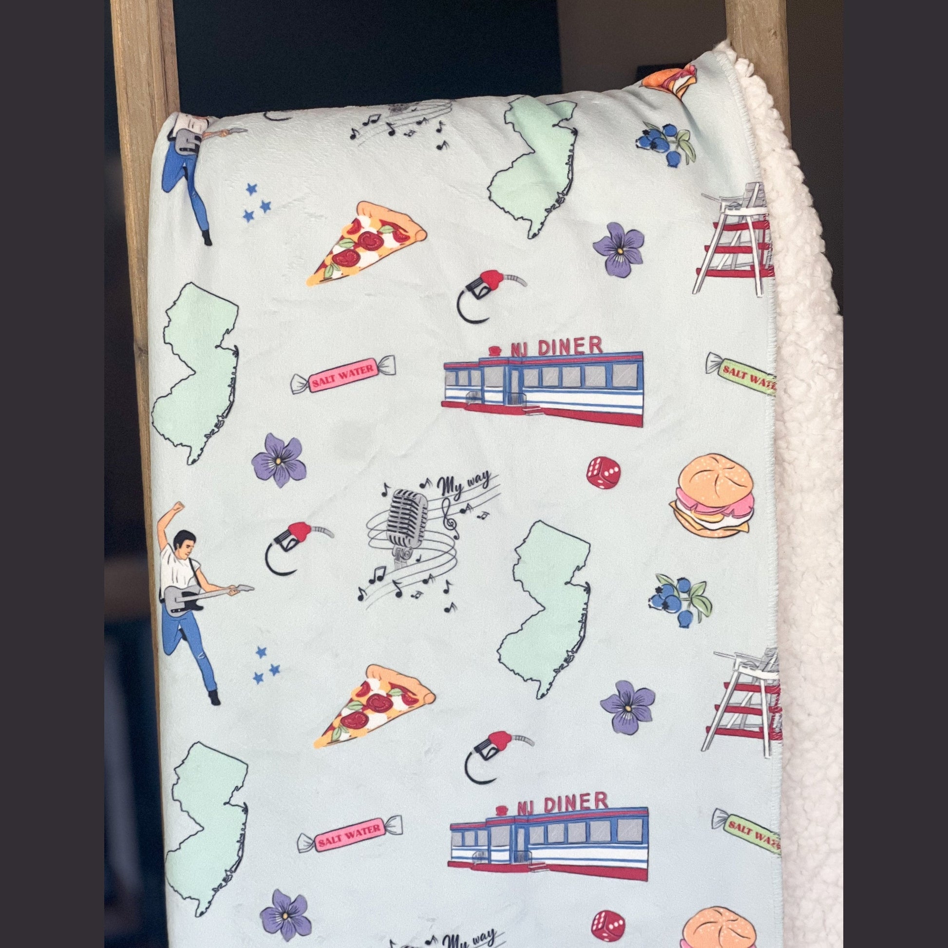 New Jersey-themed plush throw blanket, 60x80 inches, featuring a detailed map with landmarks, a rock star with his guitar, pizza, a diner, a gas pump, musical notes, flowers, and vibrant colors.