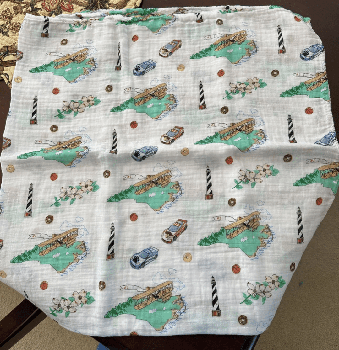 White muslin swaddle blanket with a colorful map of North Carolina, featuring landmarks and playful illustrations.