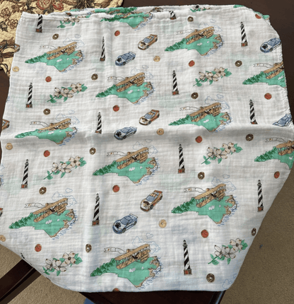 White muslin swaddle blanket with a colorful map of North Carolina, featuring landmarks and playful illustrations.
