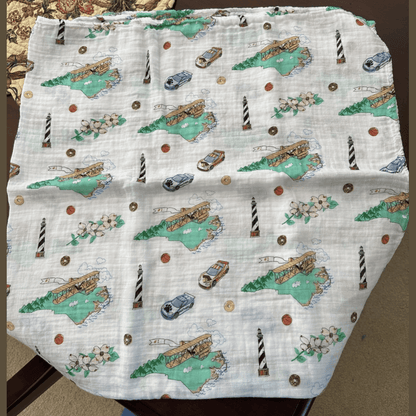 White muslin swaddle blanket with a colorful map of North Carolina, featuring landmarks and playful illustrations.