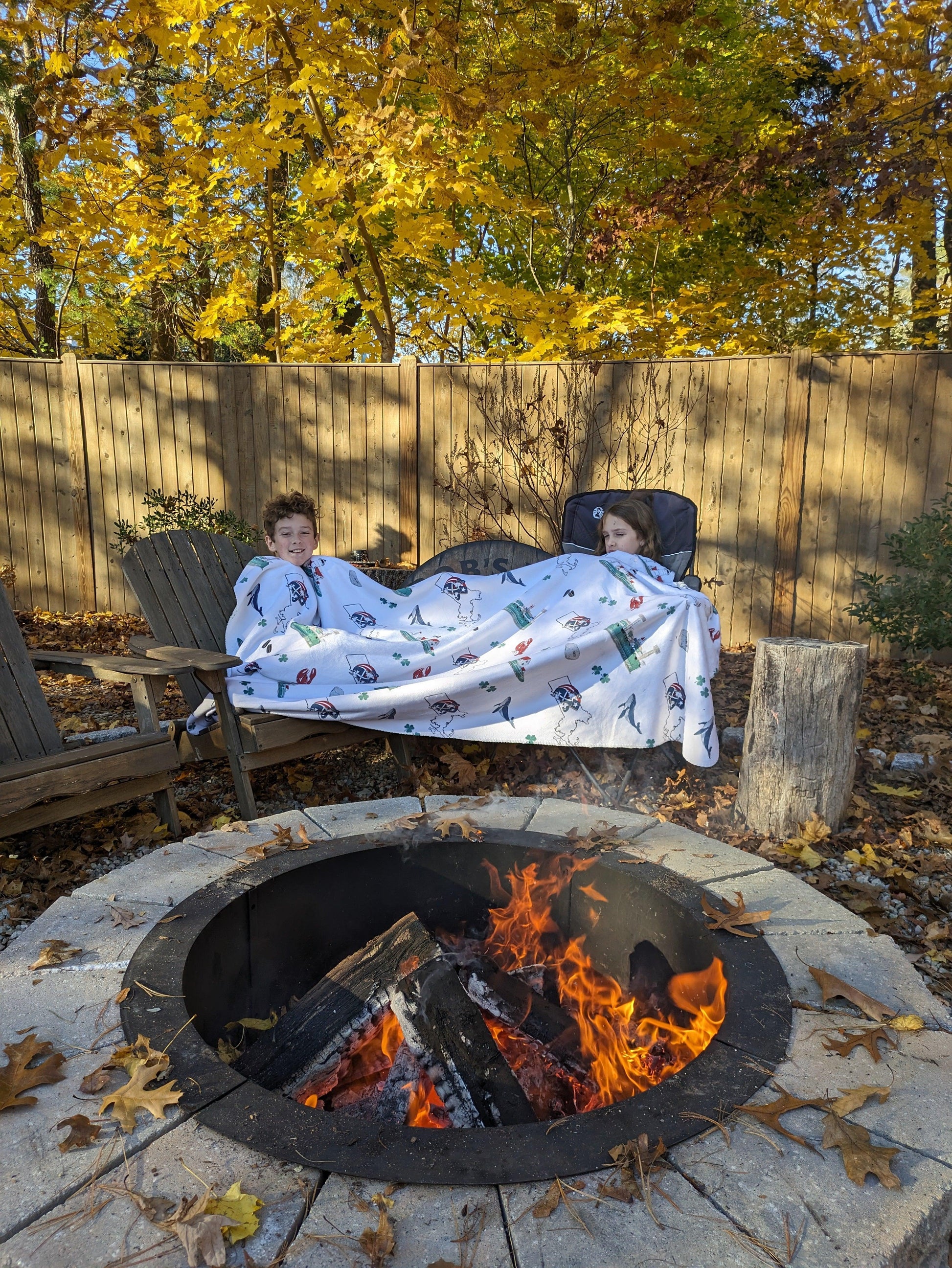A boy and girl cozying up by a firepit in front of a fence on a cool Autumn day both sharing a large Massachusetts-themed plush blanket  bundle with featuring state icons and vibrant colors.