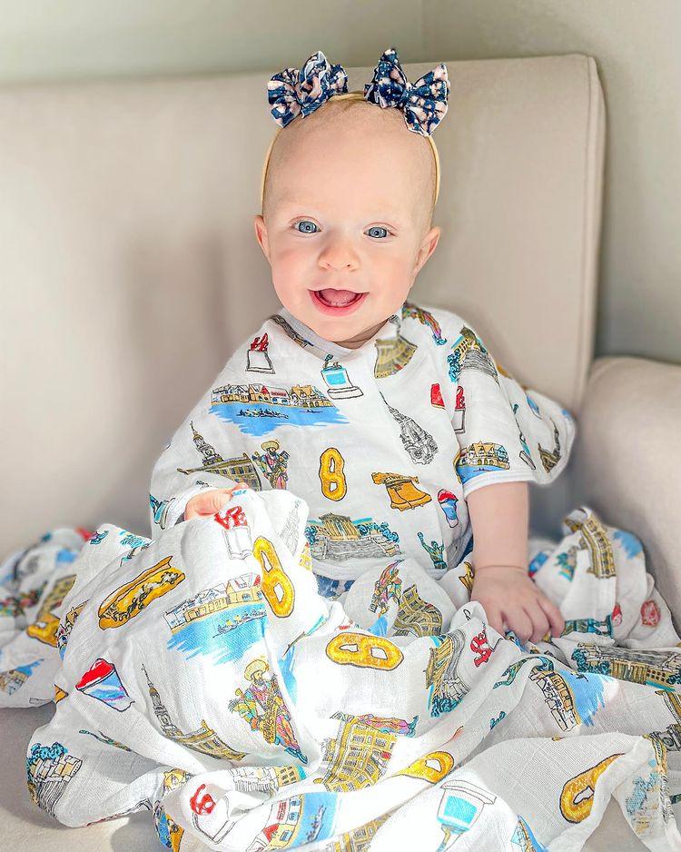 A smiling baby girl wearing bows and wrapped in a vibrant Philadelphia-themed baby bib and swaddle gift set.  It includes famous foods, landmarks, and cultural icons of Philadelphia