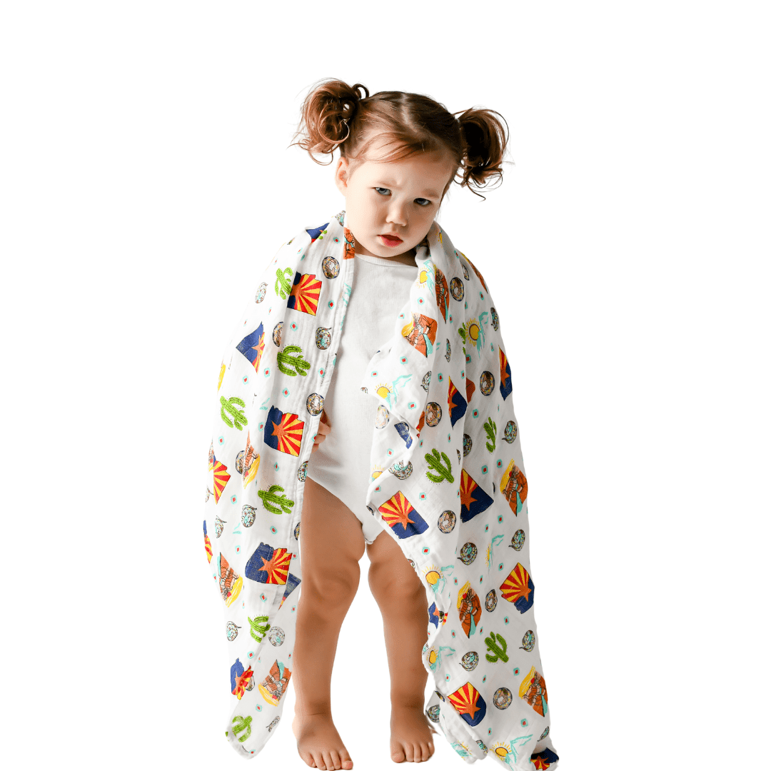 White muslin swaddle blanket with a colorful Arizona-themed print, featuring cacti, mountains, and desert animals.