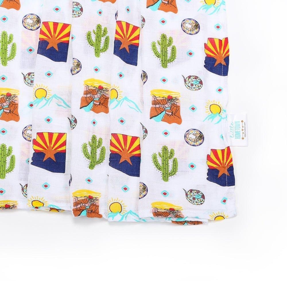 Baby swaddled in a white muslin blanket with colorful Arizona-themed illustrations, including cacti and desert animals.