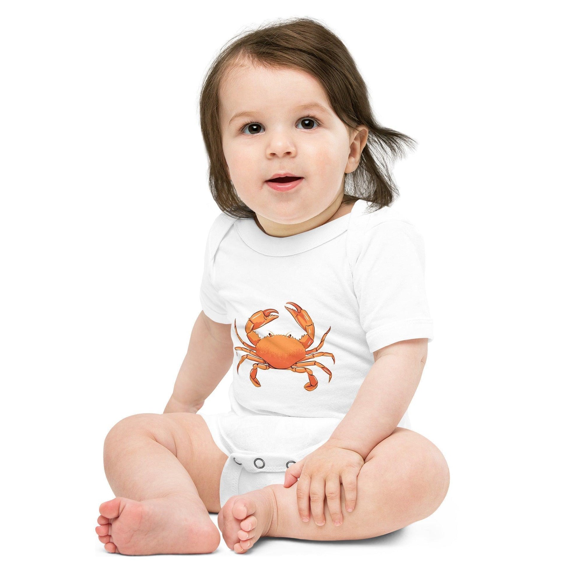 A baby with long brown hair wearing a white baby onesies with a light red crab printed on it
