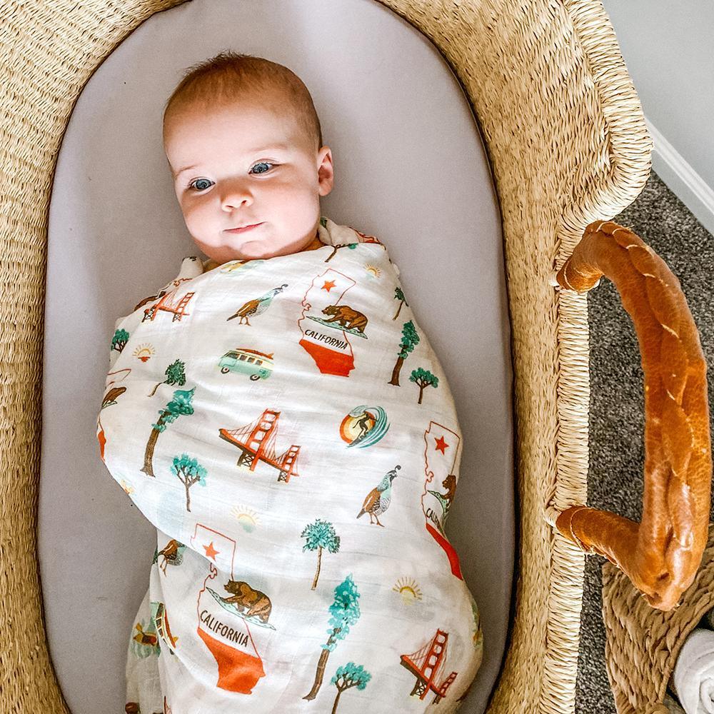 White muslin swaddle blanket with a California-themed print featuring landmarks, icons, and text in soft pastel colors.