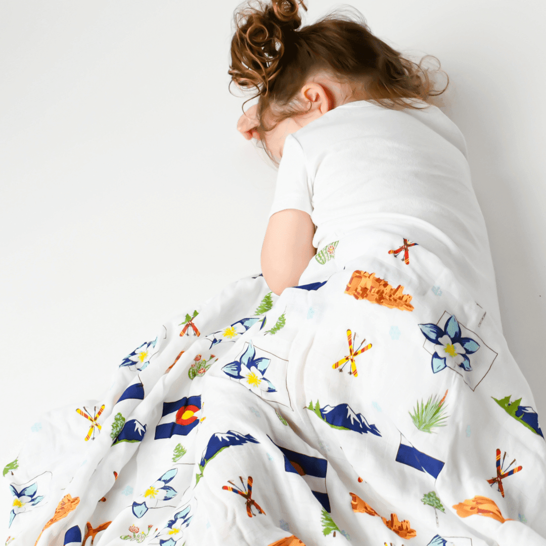 White muslin swaddle blanket with colorful Colorado-themed illustrations, including mountains, trees, and wildlife.