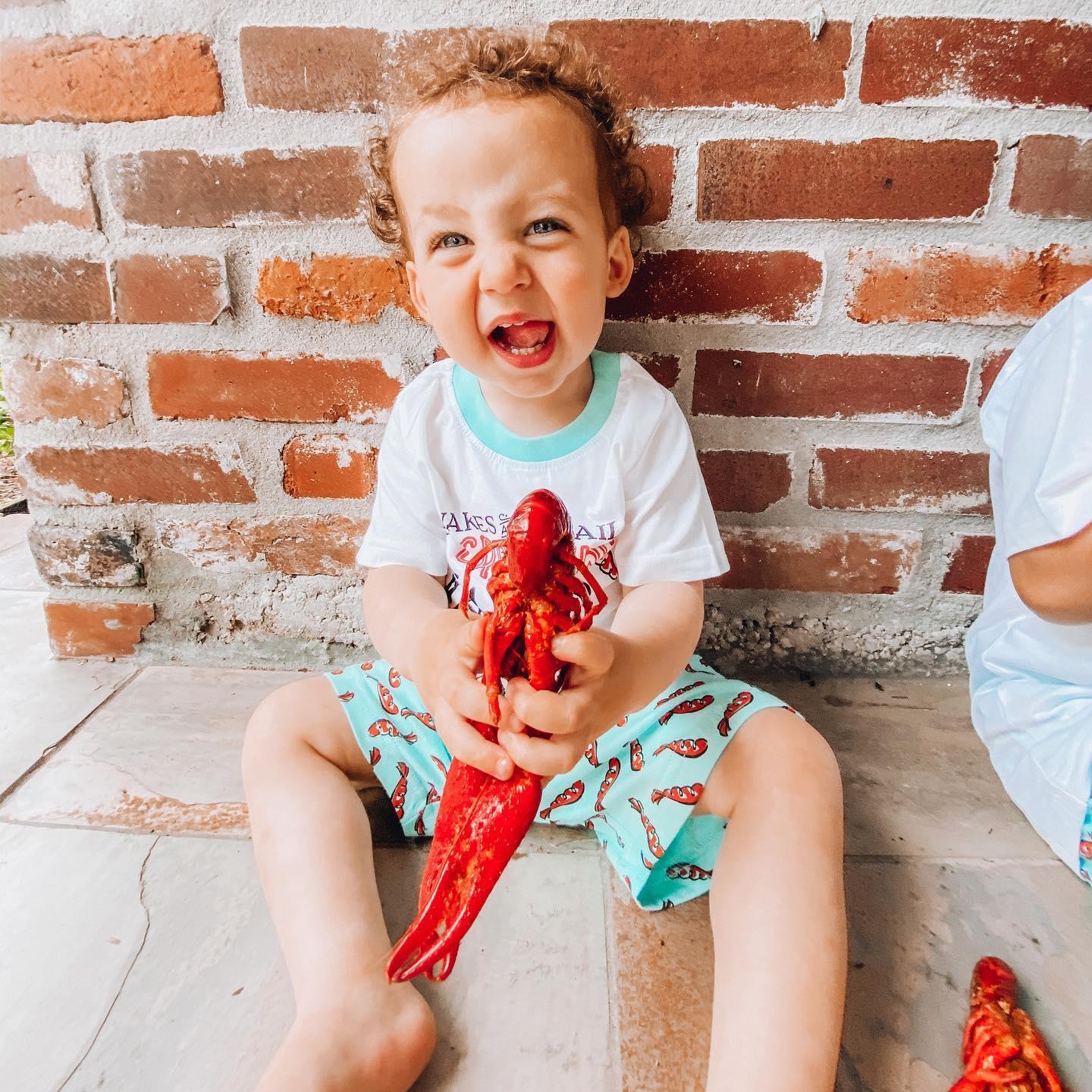 Boy wearing blue pajamas with red crawfish pattern, smiling and holding a toy, sitting on a white bed.