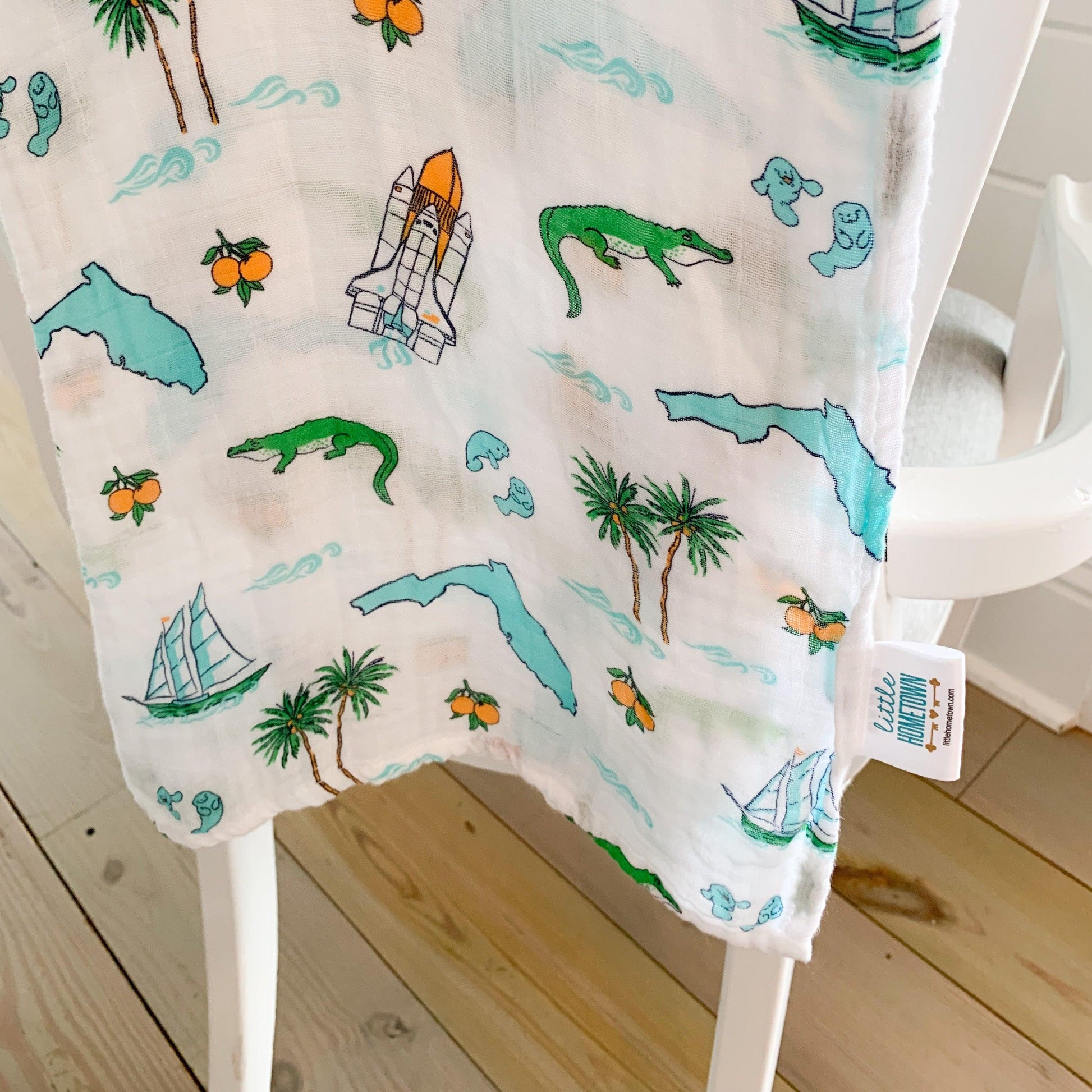 Florida-themed baby muslin swaddle blanket with colorful illustrations of palm trees, flamingos, and oranges.