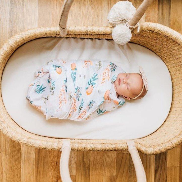 Baby swaddled in a white muslin blanket with "Georgia Girl" text and peach illustrations, lying on a soft surface.