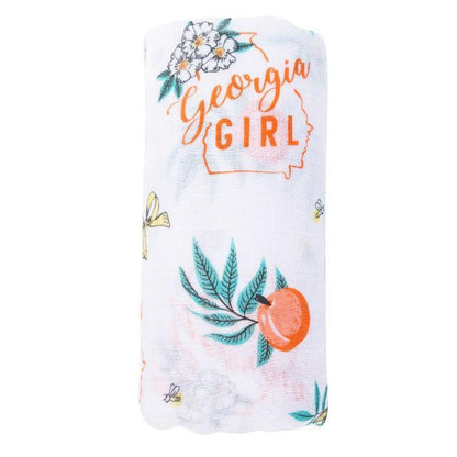 Baby swaddled in a white muslin blanket with "Georgia Girl" text and peach illustrations, lying on a soft surface.