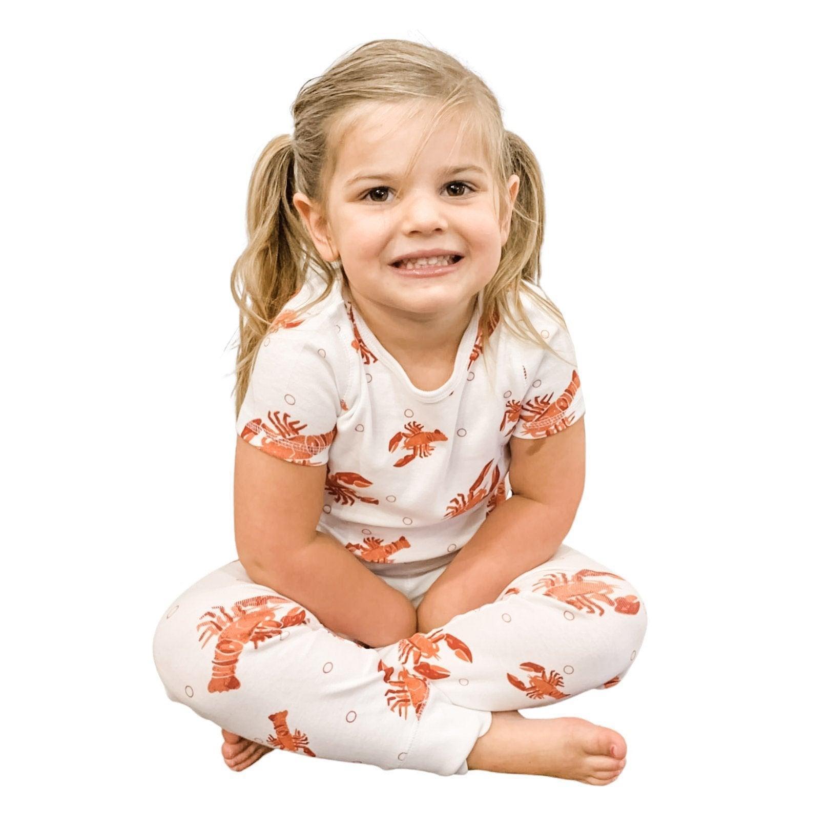 Toddler wearing white pajamas with red lobsters and blue crawfish, smiling and sitting on a white bed.