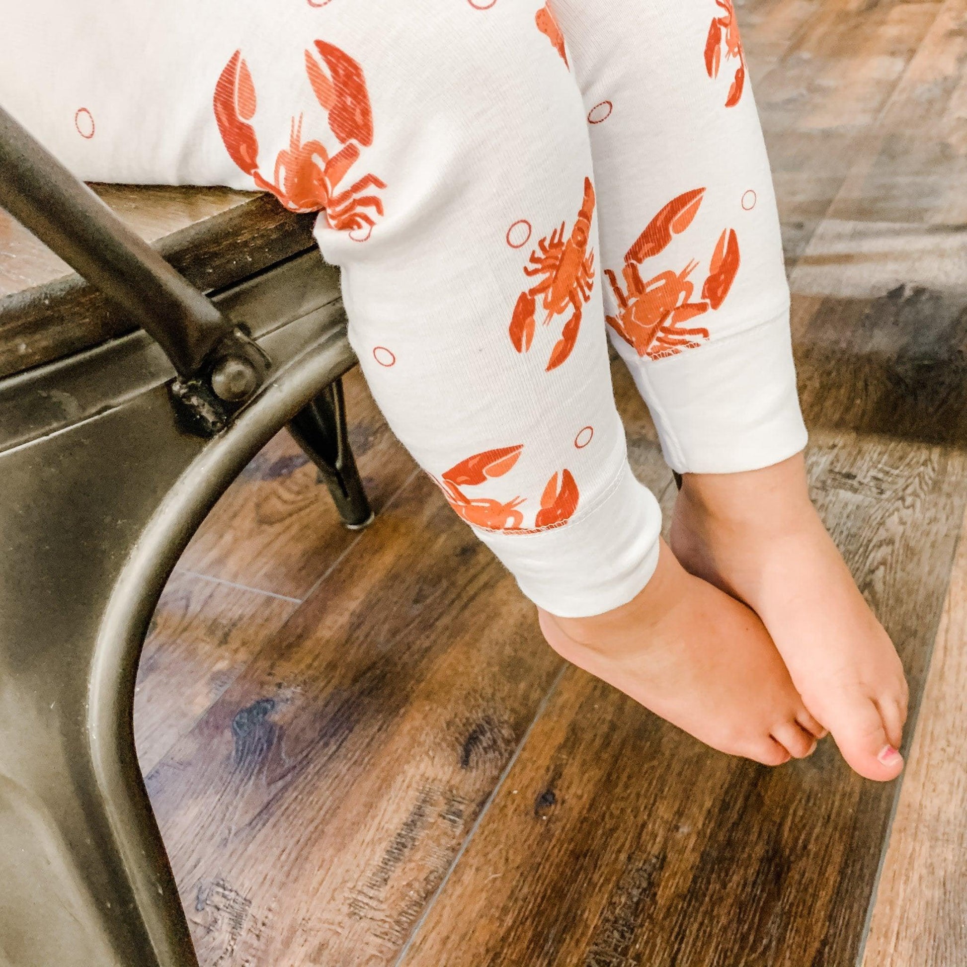 Toddler wearing white pajamas with red lobsters and crawfish, smiling and sitting on a white bed.