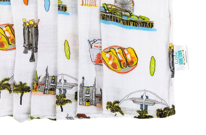 Los Angeles-themed baby muslin swaddle blanket with iconic landmarks and pastel illustrations on a white background.