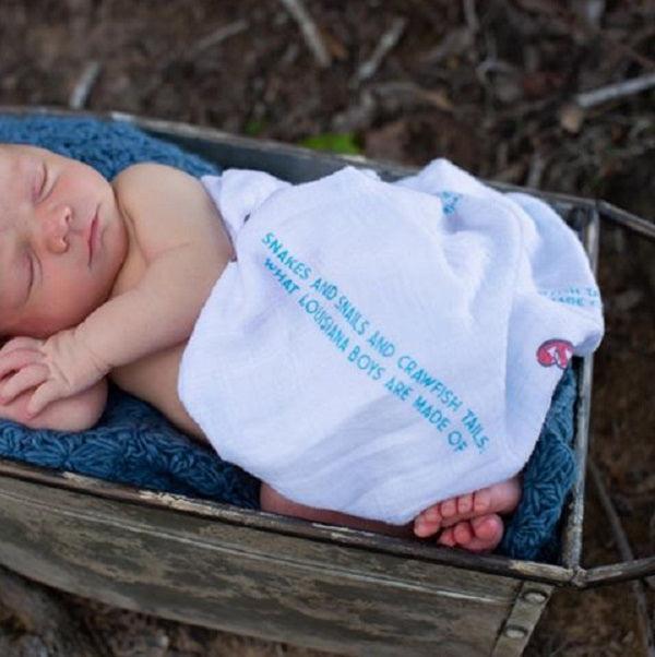 Louisiana-themed baby muslin swaddle blanket with state icons like pelican, magnolia, and crawfish on a white background.