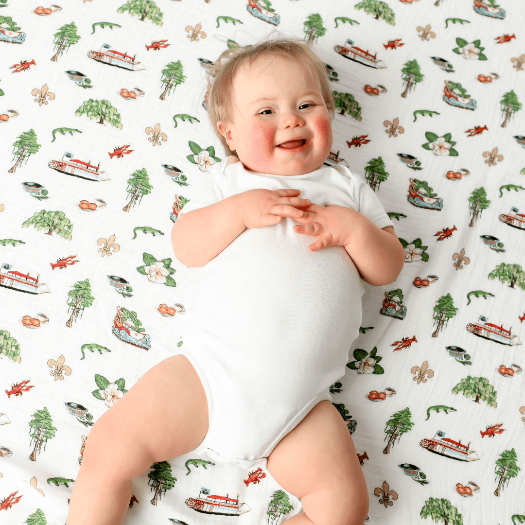 Louisiana-themed baby muslin swaddle with state icons like pelicans, magnolias, and crawfish on a white background.