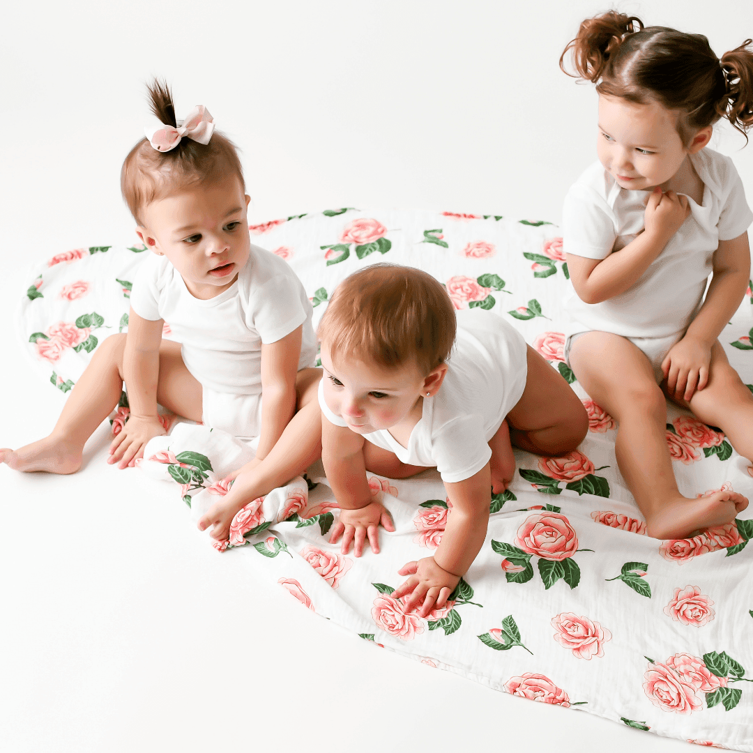 Soft muslin baby swaddle blanket with delicate pink camellia flowers and green leaves on a white background.