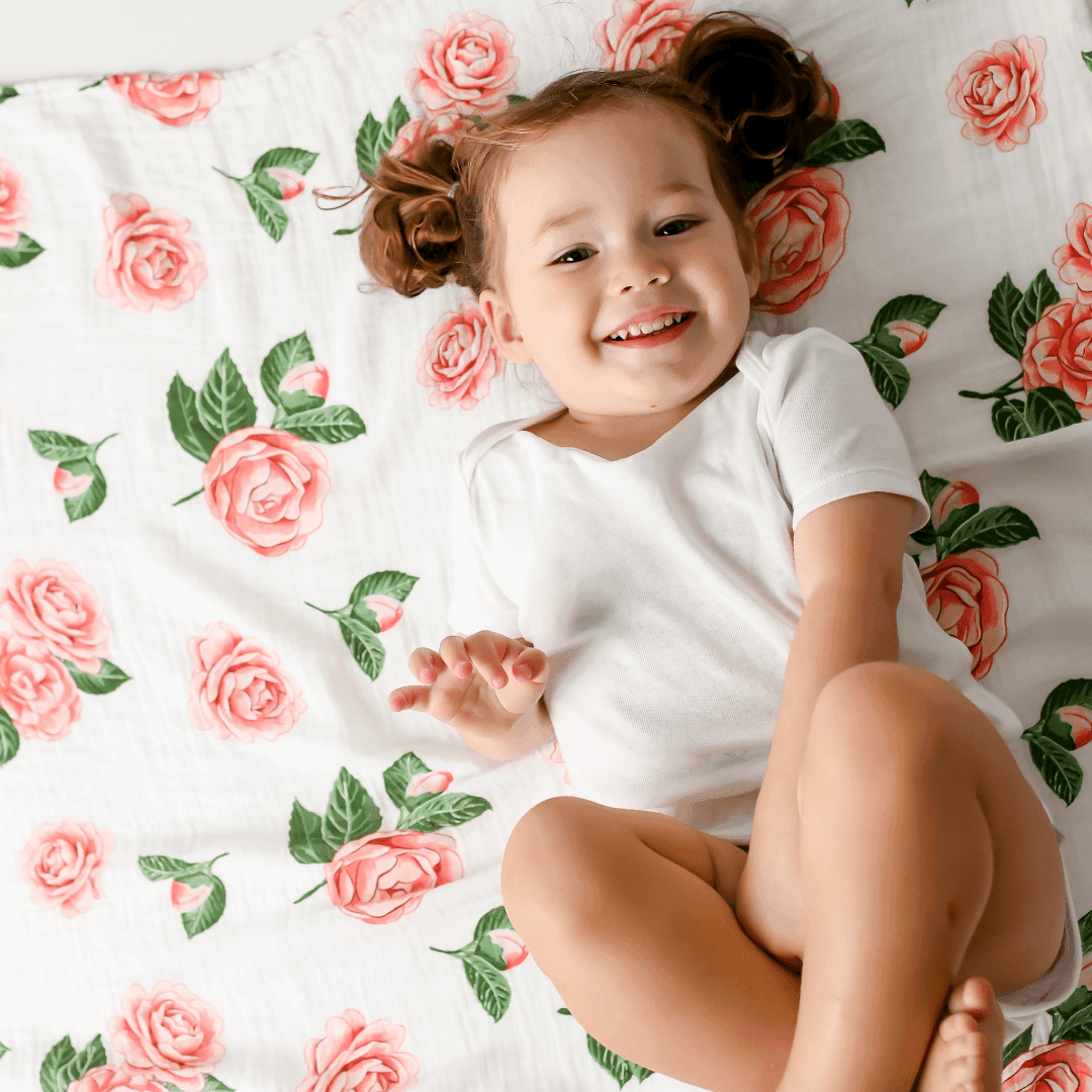 Soft muslin baby swaddle blanket with delicate pink camellia flowers and green leaves on a white background.