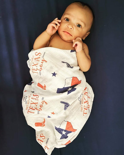 White muslin swaddle blanket with blue Texas-themed illustrations, including the state outline, cowboy boots, and stars.